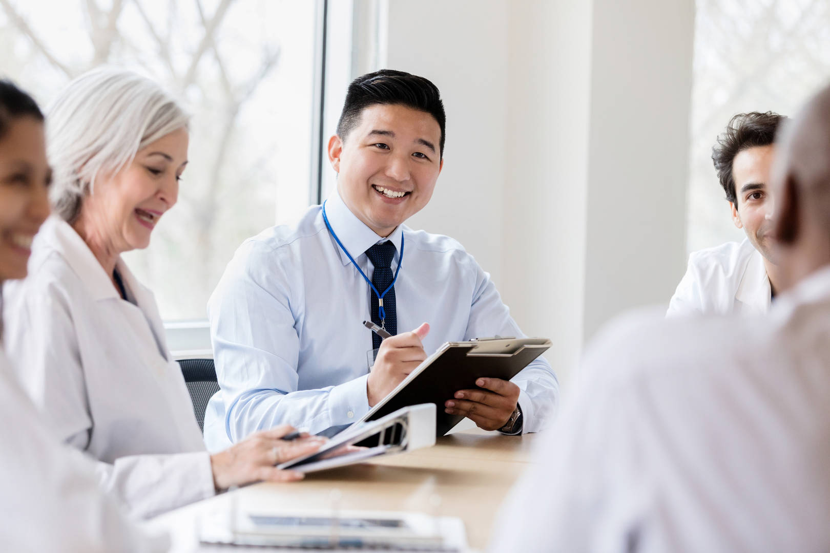 Young Asian male hospital administrator takes notes during meeting with healthcare professionals. He is talking with a male doctor.