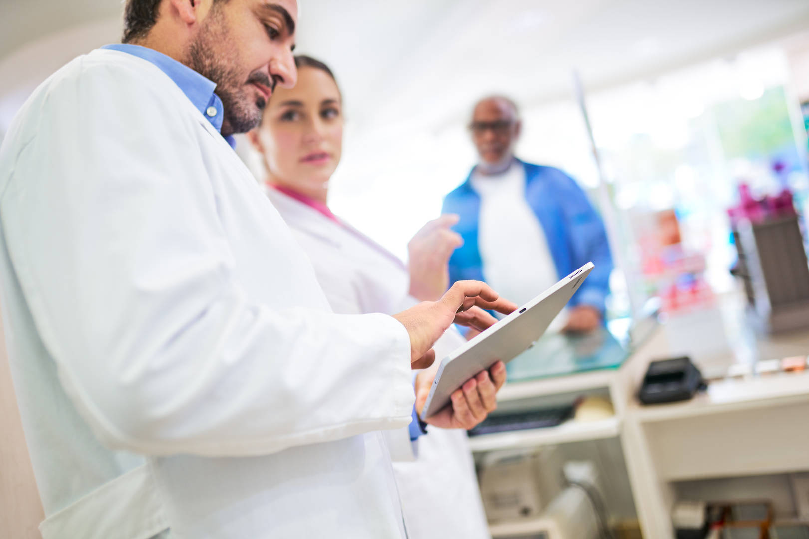 Two pharmacists working on a tablet while a patient is at the counter. 