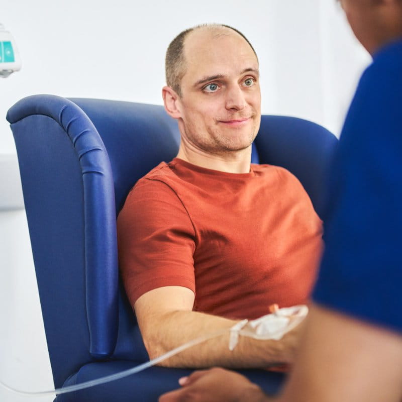 A male patient in an infusion chair speaking to a nurse.